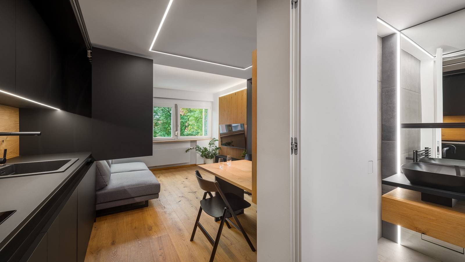 Minimalist studio apartment with a kitchen, a wooden dining table, a grey sofa and a wall-mounted TV