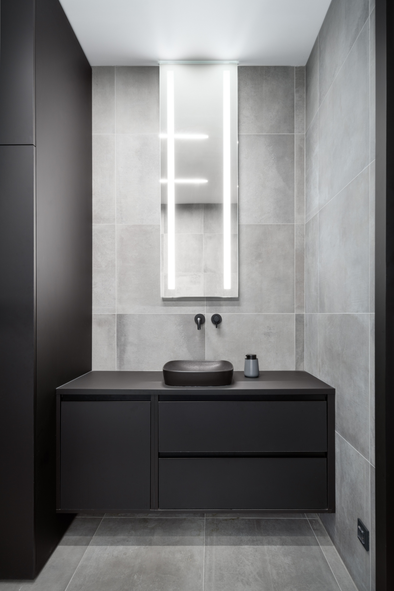 Minimalist bathroom with grey tiles, black furniture, and an illuminated mirror