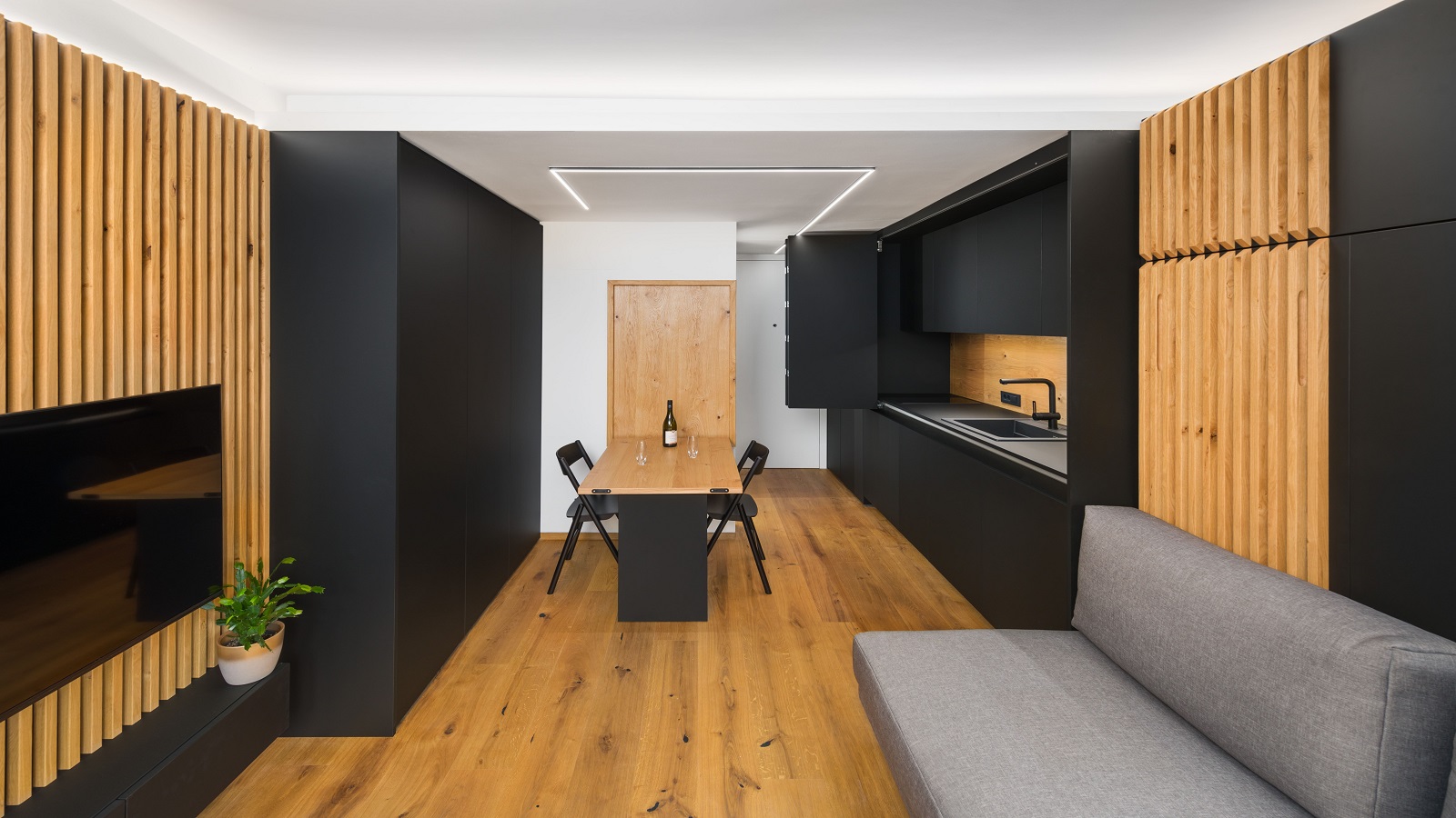 Studio apartment with wooden floors, black cabinets, a dining table, a sofa and a wall mounted TV.