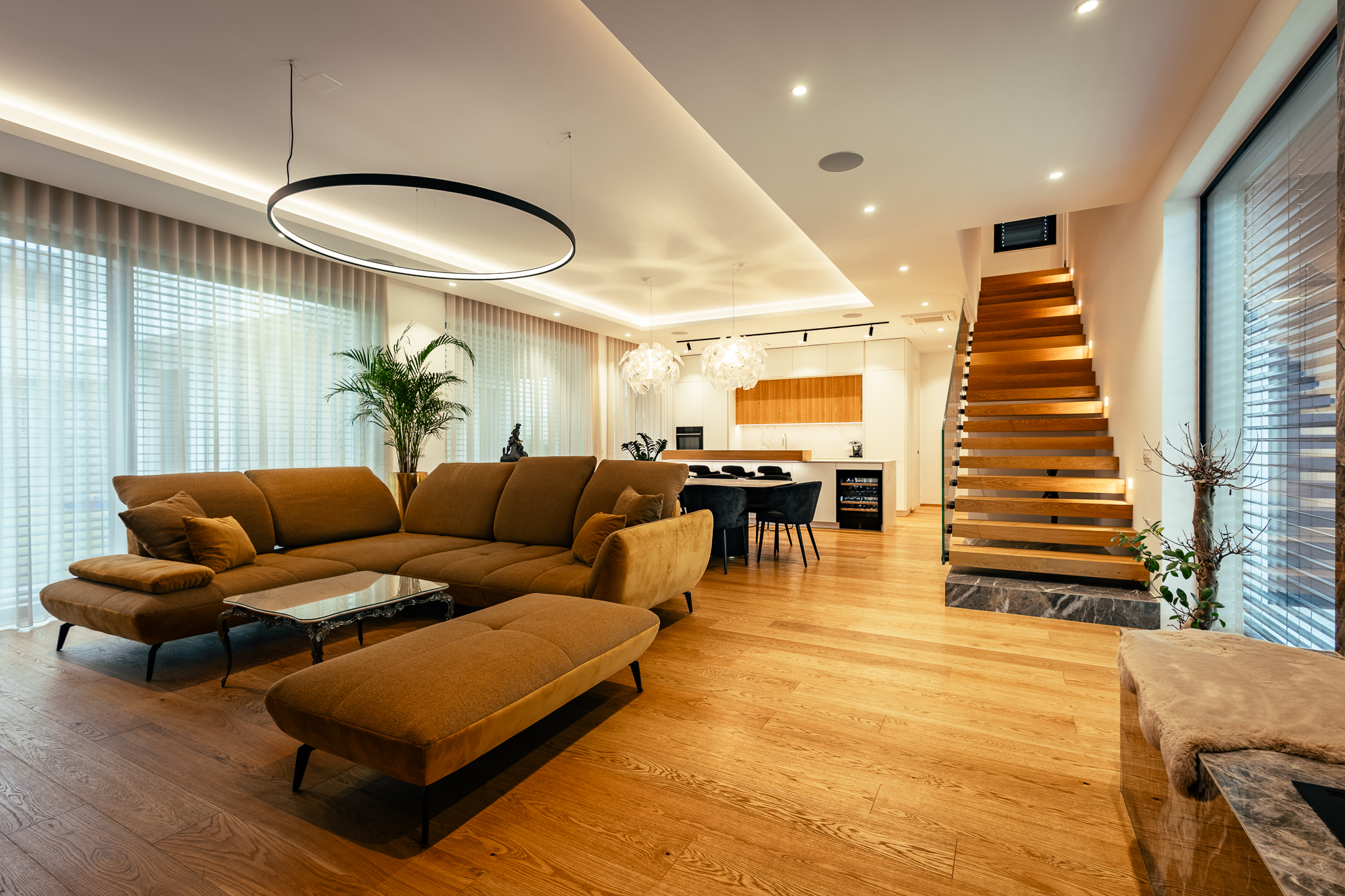 Dnevna soba z lesenim parketom, belimi stenami, jedilnico, kuhinjo, stopniščem in rjavo sedežno garnituro v sredini katere je klubska mizica