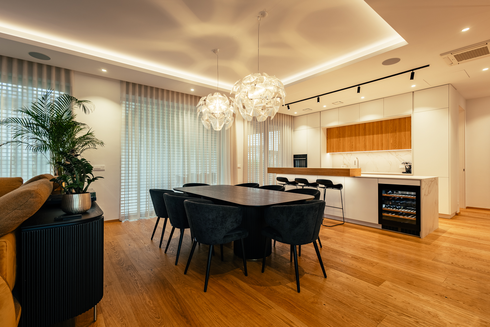 Jedilnica v luksuzni vili, s črno jedilno mizo, svtetlo-leseno kuhinjo in lestenci