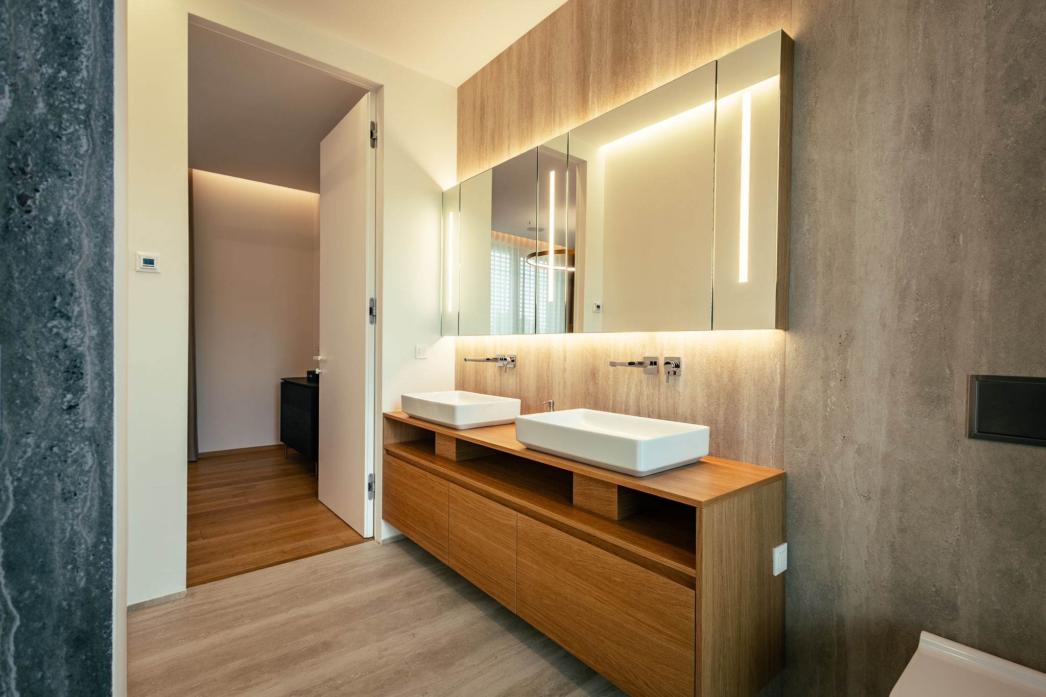 Kopalnica, ki ima sive stene, z dvema umivalnikoma, lesenimi policami in steklenimi, osvetljenimi omaricami