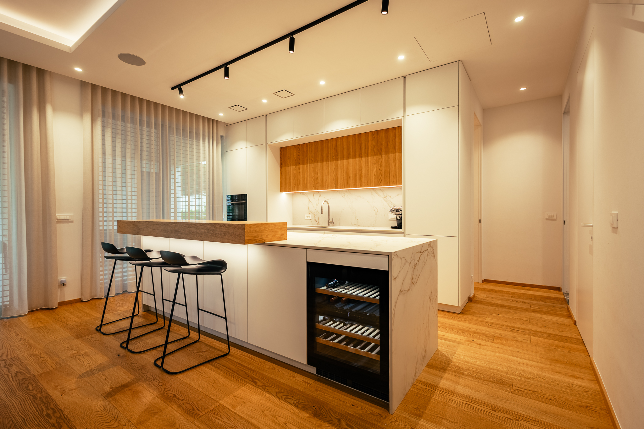 Kuhinja luksuzne vile z belim, marmornatim kuhinjskim otokom, za katerim so postavljeni trije, črni barski stoli, v njem pa je vgrajen vinski hladilnik