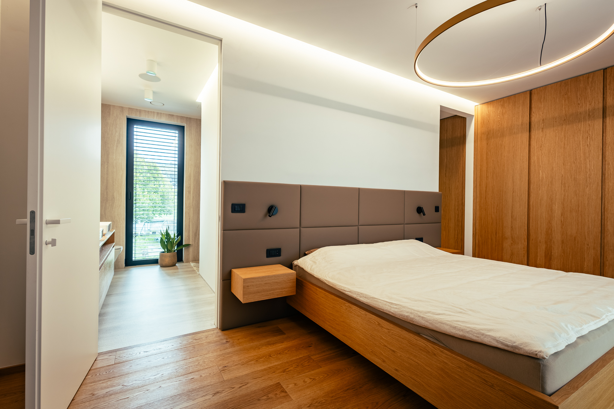 Svetla spalnica s posteljo, lesenim parketom, leseno omaro in vidnim prehodom v privatno kopalnico